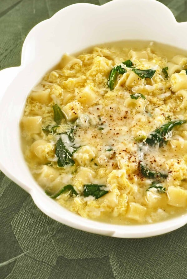 A white bowl of Italian egg drop soup with spinach and ditalini, topped with grated Parmesan cheese and a sprinkle of nutmeg.