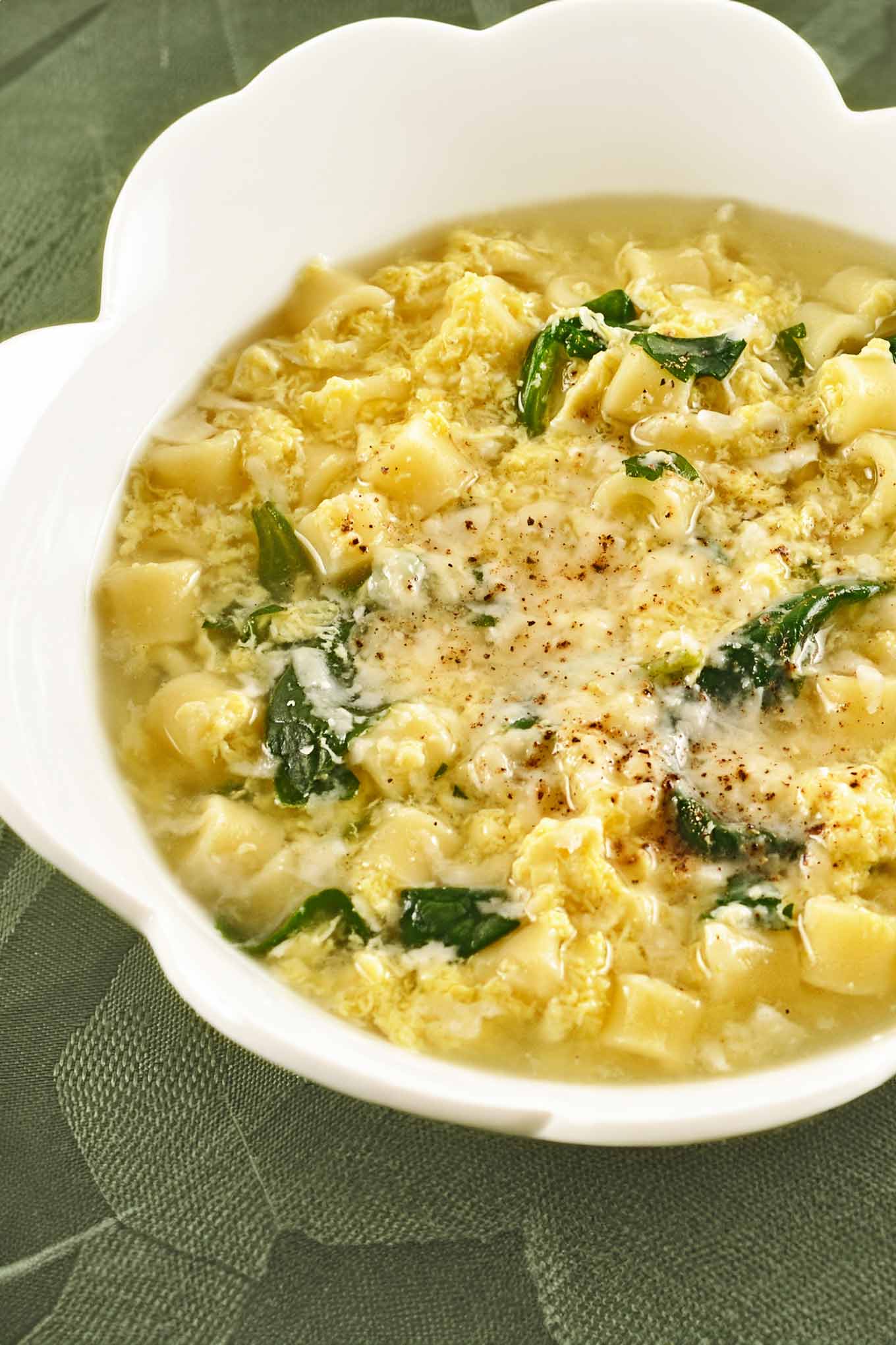 A white bowl of Italian egg drop soup with spinach and ditalini, topped with grated Parmesan cheese and a sprinkle of nutmeg.