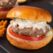 A serving board with a grilled lamb burger on a brioche bun with tzatziki sauce and slices of onion and tomato.