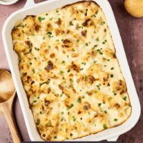 Golden-browned cottage cheese potato casserole in a white baking dish, topped with melted cheese and chopped chives, on a rustic surface with whole potatoes and a wooden spoon nearby.