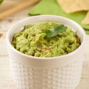 Chunky guacamole in a white ramekin, garnished with a sprig of cilantro, with tortilla chips and lime in the background.