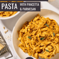 Two bowls of creamy pumpkin pasta with pancetta and sage served in white bowls, a glass of white wine and cheese grater in the background.