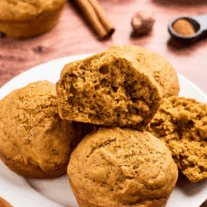 Soft, golden pumpkin spice muffins on a plate with cinnamon sticks in the background, and one muffin cut open to show the tender, moist crumb.