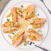 An overhead view of a round white plate filled with baked stuffed colossal shrimp with seafood forks and lemon slices for garnish.