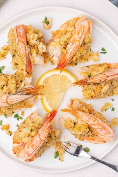 An overhead view of a round white plate filled with baked stuffed colossal shrimp with seafood forks and lemon slices for garnish.