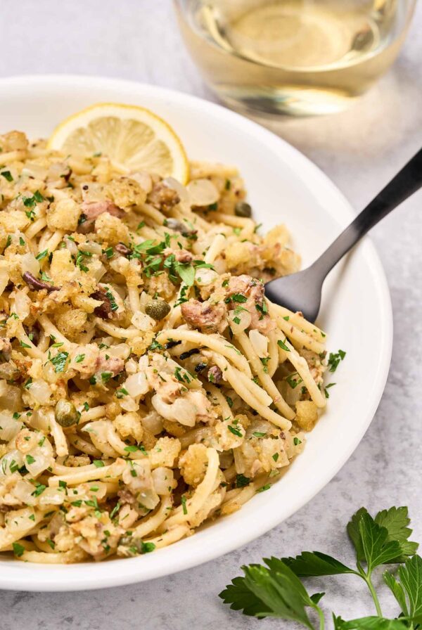 A serving of sardine pasta with toasted garlic breadcrumbs in a white bowl with a black ford and a lemon wedge, fresh parsley, and a glass of white wine on the side.