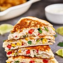 Chicken and cheese quesadillas cut into wedges and stacked on a cookie sheet along with lime slices and a bowl of sour cream.