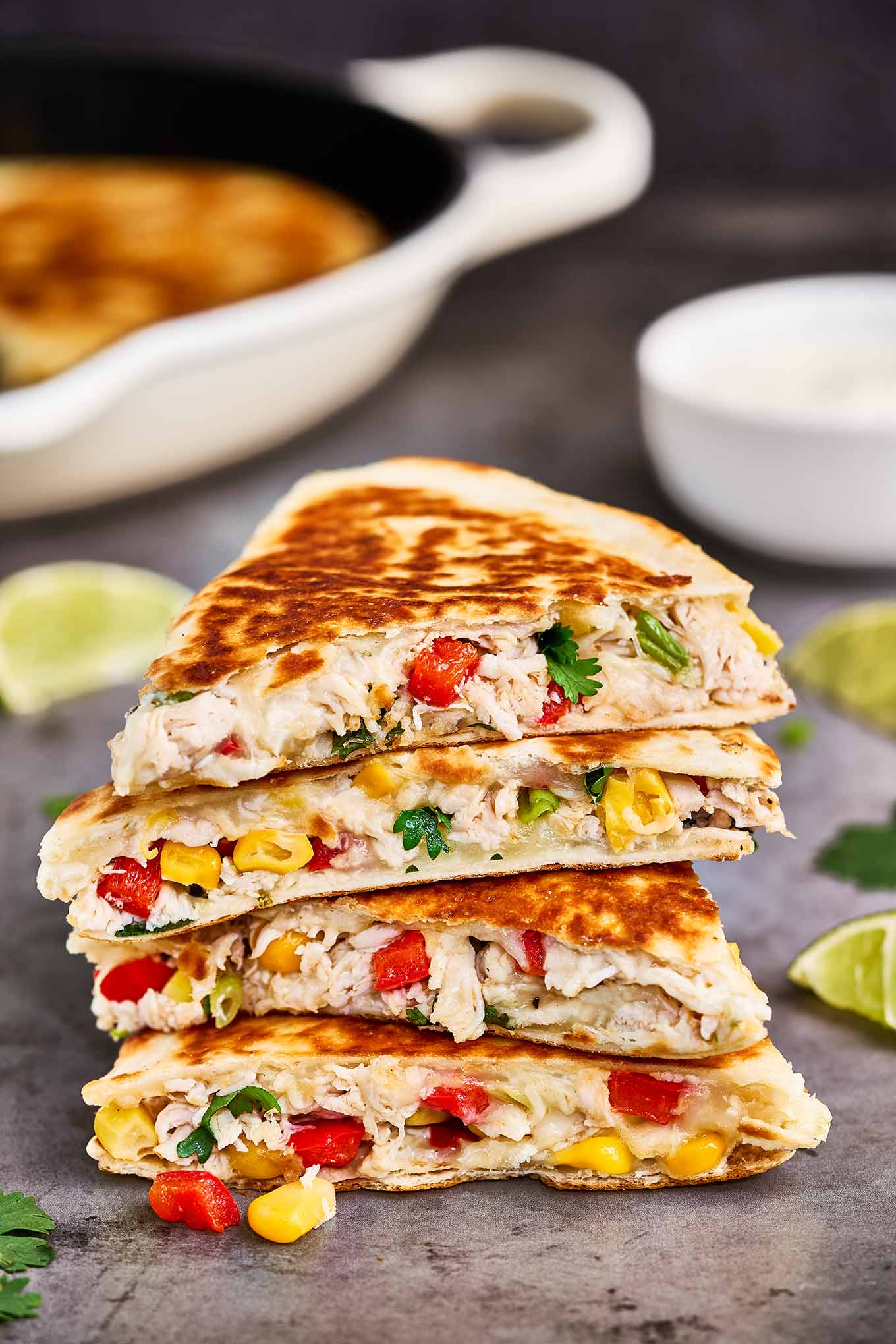 Chicken and cheese quesadillas cut into wedges and stacked on a cookie sheet alongside lime wedges, cilantro, and a dish of sour cream.