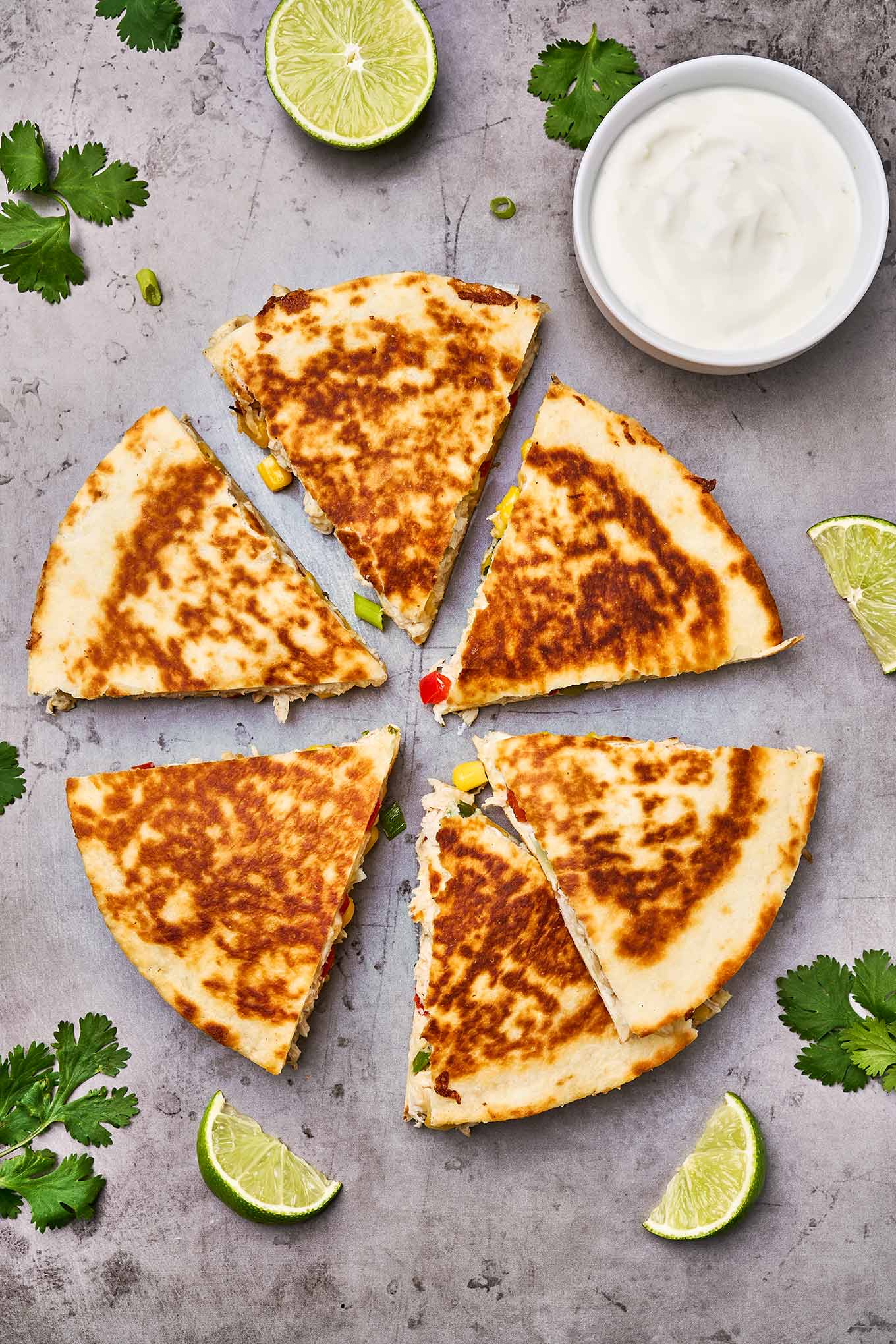 An overhead view of chicken and cheese quesadillas cut into wedges surrounded by lime slices, fresh cilantro, and sour cream for dipping.
