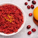 A serving bowl of fresh cranberry orange relish garnished with orange zest, on a gray background with scattered cranberries to the right.