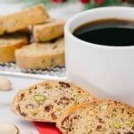 Cranberry pistachio biscotti in front of a cup of coffee, with more biscotti on a cooling rack in the background.