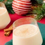 A cinnamon-dusted glass of spiked eggnog on a green cocktail napkin with cinnamon sticks and Christmas decor in the background.