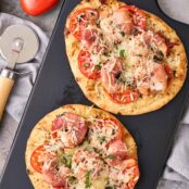 Two naan pizzas topped with prosciutto-wrapped shrimp, tomatoes, basil, and mozzarella on a black board with a pizza cutter.