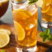 An ice-filled glass of boubon-spiked iced tea with lemons and mint, with more glasses and the pitcher in the background.