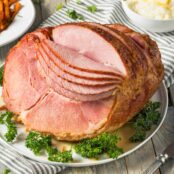 A large, spiral-sliced, glazed ham on a serving platter, surrounded by parsley garnish, mashed potatoes and a striped tea towel in the background.
