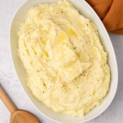 An oval serving bowl of creamy mashed potatoes with a melting pat of butter in the middle, a wooden spoon and linen napkin on either side of the bowl.