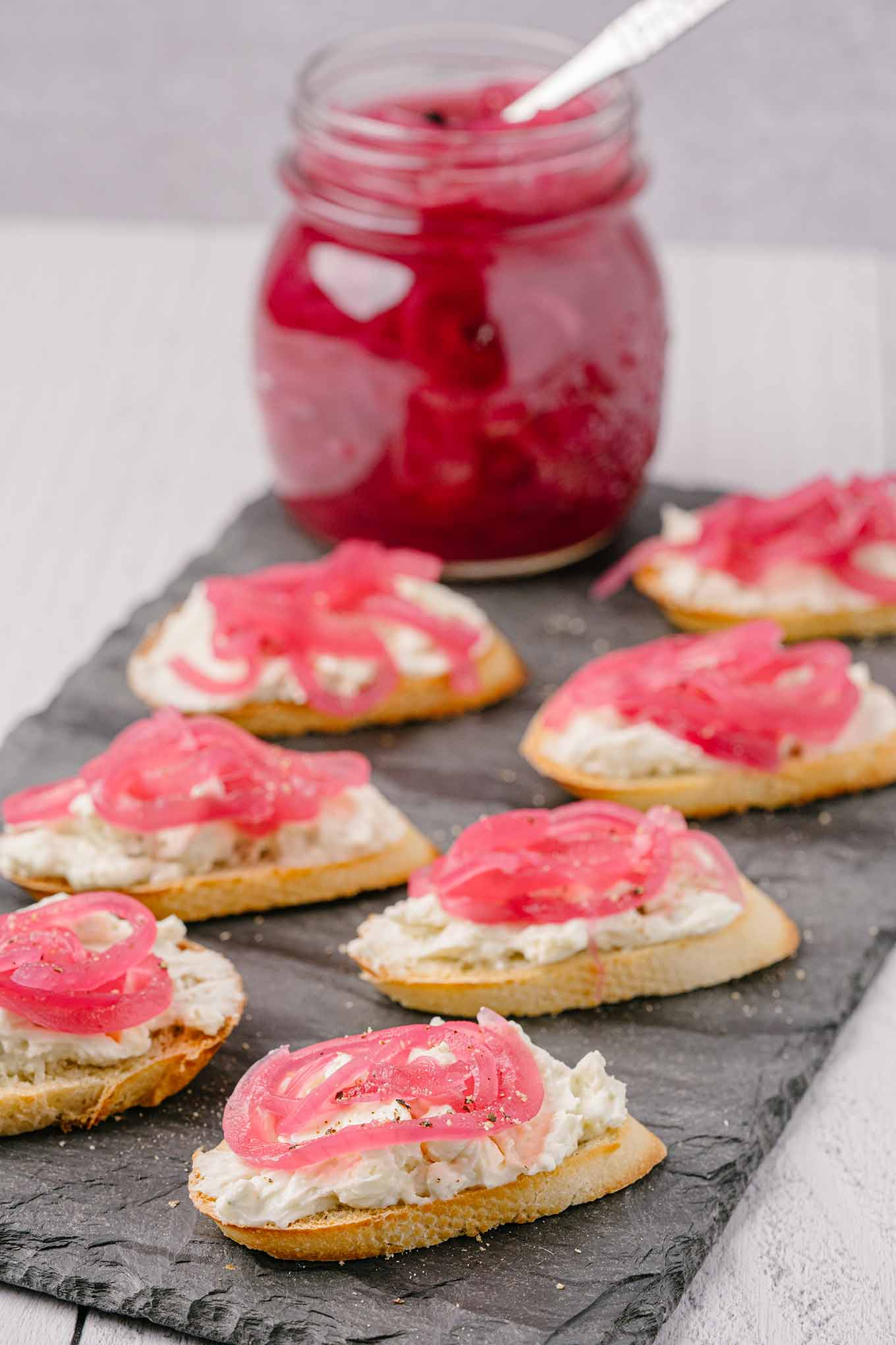 Six slices of toasted baguette topped with a white creamy spread and vibrant pink pickled onions, arranged on a dark rectangular platter.