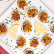 Clams Casino topped with bacon and arranged on an appetizer serving plate with lemon wedges and parsley garnish.