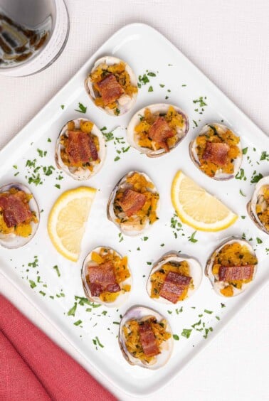 Clams Casino topped with bacon and arranged on an appetizer serving plate with lemon wedges and parsley garnish.