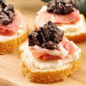 Three crostini topped with whipped ricotta, prosciutto, and port wine fig compote arranged on a wooden cutting board.