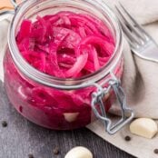 A glass canning jar filled with pickled red onions, surrounded by garlic cloves and black peppercorns on a dark wooden surface.