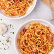 Two pasta bowls filled with linguine tossed with calamari (squid) in marinara sauce, garnished with parsley, and surrounded by a bulb of garlic, and an olivewood spoon.