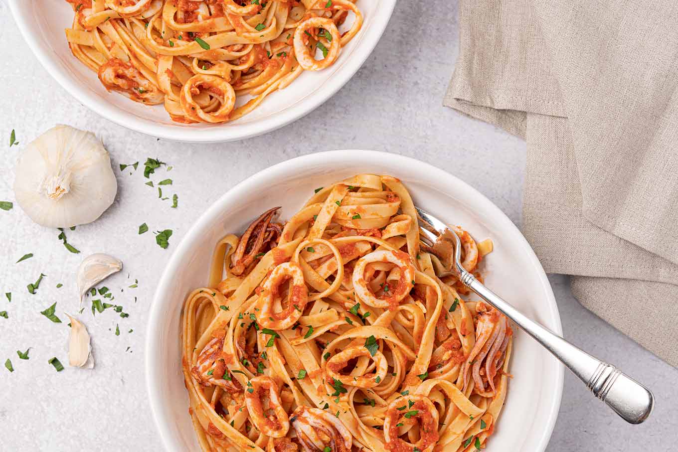 Two bowls of linguine with squid in marinara sauce, a bulb of garlic, scattered parsley, and a linen napkin.