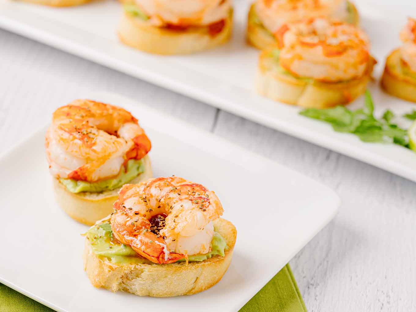 A serving of two shrimp crostini on a white appetizer plate with the serving platter in the background.