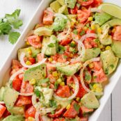 A serving platter of avocado chunks, tomatoes, corn kernels, sliced onion, and cilantro, tossed in creamy lime dressing and garnished with lime wedges.