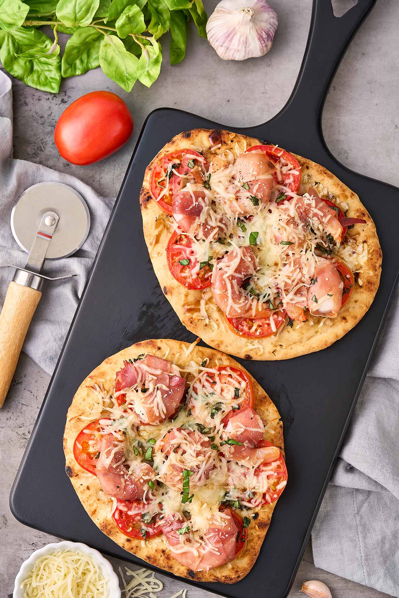 A black cutting board with two naan pizzas topped with prosciutto-wrapped shrimp, sliced tomatoes, fresh basil, and shredded mozzarella.