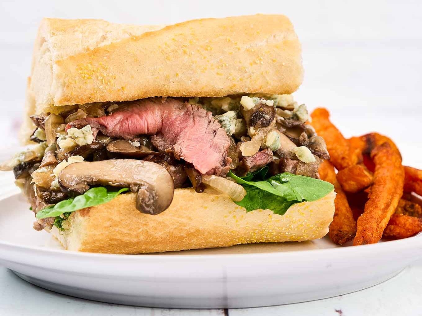 A single portion of a grilled ribeye steak sandwich with blue cheese on a white oval plate with sweet potato fries on the side.