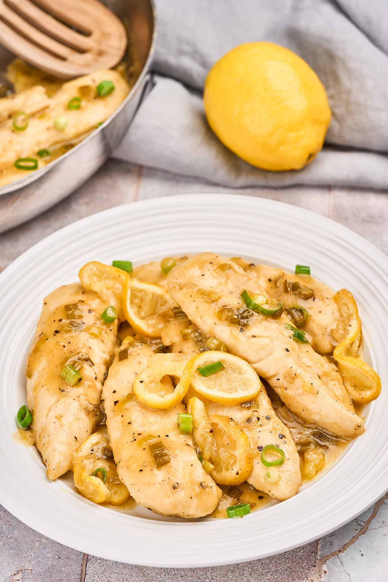 A shallow white serving bowl filled with lemon pepper chicken tenders in pan sauce, garnished with lemon slices and chopped scallions.