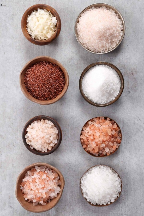 Eight small bowls filled with different types of salt on a gray background.