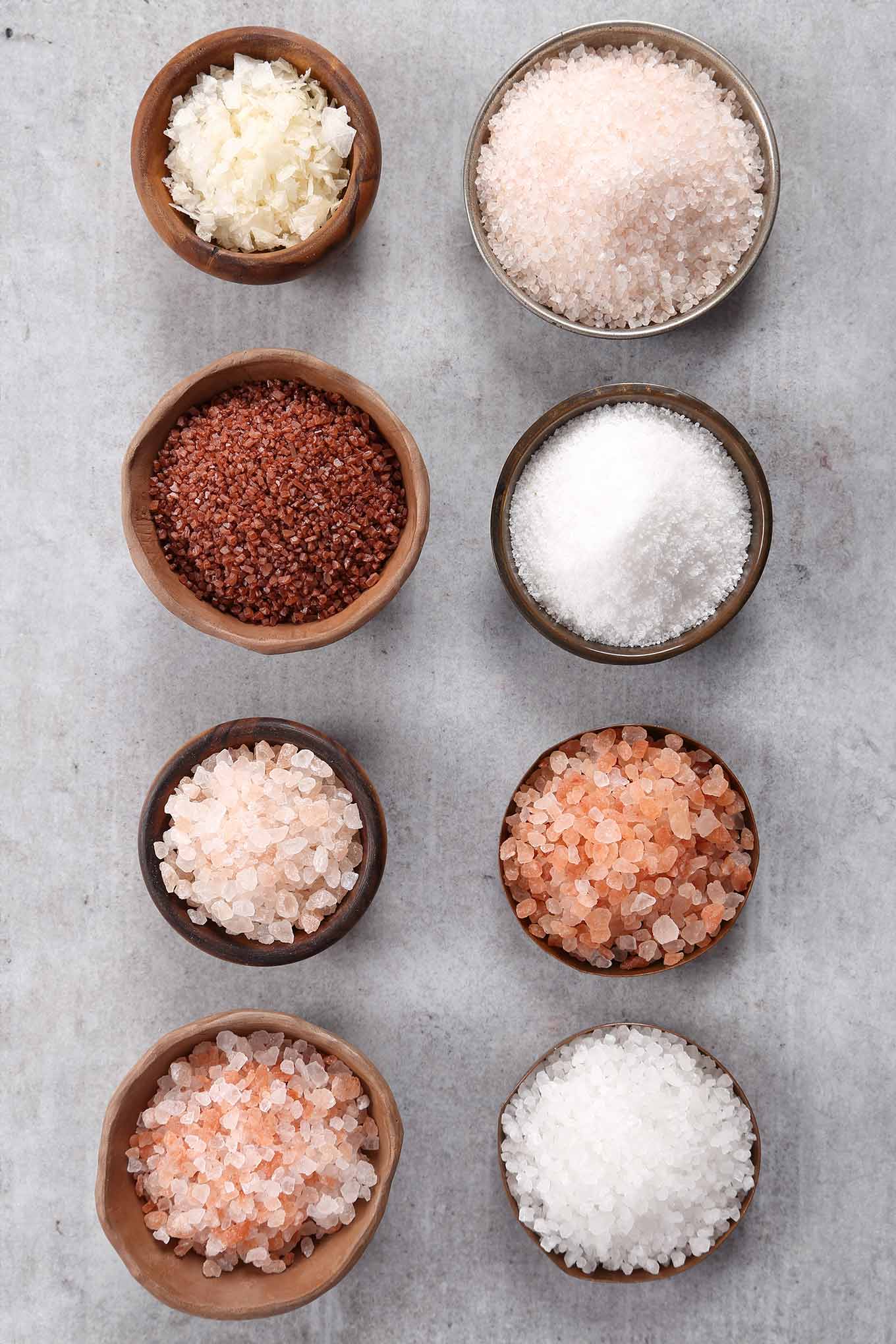 A variety of different salts in small bowls including kosher salt, Himalayan pink salt, fleur de sel, table salt, and red Hawaiian sea salt.