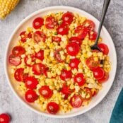 Fresh corn and cherry tomato salad in a serving bowl with a serving spoon, surrounded by a linen napkin, corn with the husk on and cherry tomatoes.