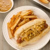 An overhead view of a plate with grilled bratwurst on a roll with sautéed cabbage and onions, a drizzle of honey mustard, and a side of French fries.
