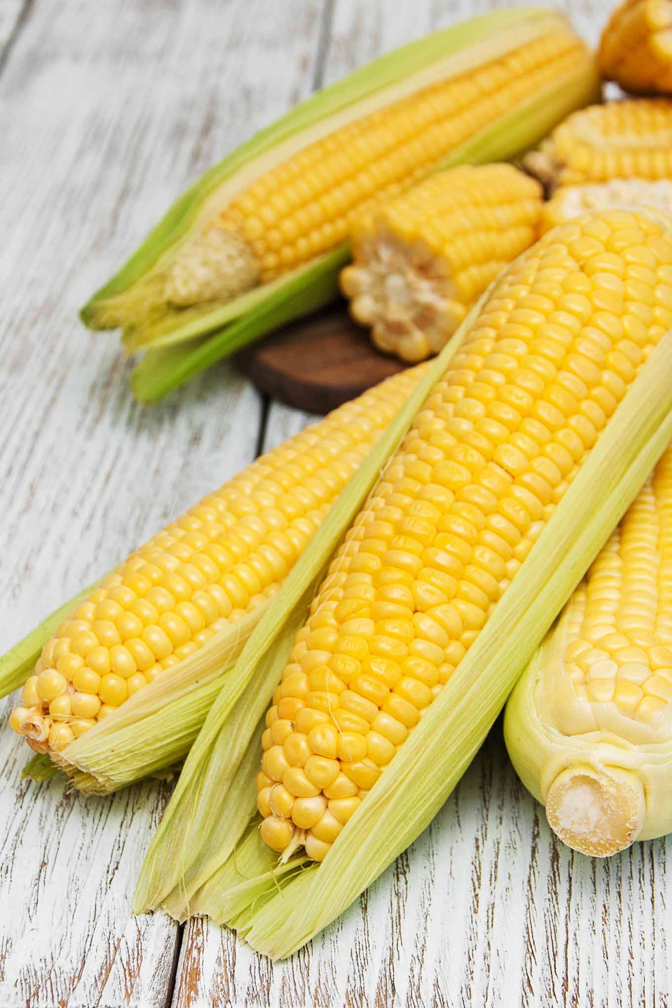 Ears of fresh corn being prepared for cooking, husks partially removed, on a gray wooden surface.