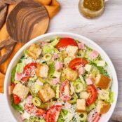 A dinner-sized serving of 1905 salad in a white bowl with croutons, grated Parmesan, olives and cubes of ham; wooden salad servers to the side.