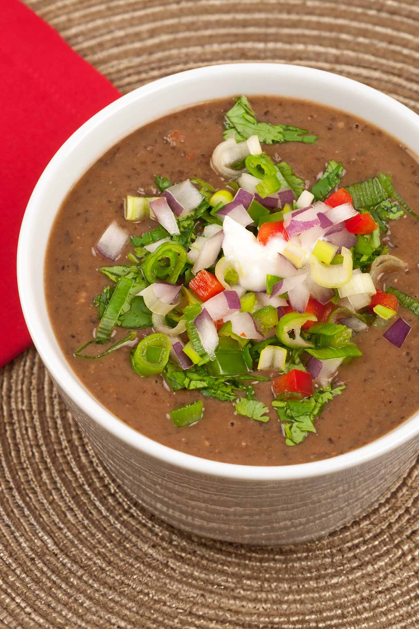 A close-up photo of a cup of black bean soup topped with diced fresh vegetables including jalapeno and red bell peppers, red onion, scallions, and cilantro on a rattan placemat.