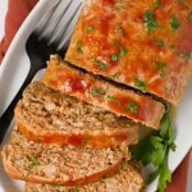 Overhead view of glazed chicken meatloaf with slices cut, parsley garnish, black fork, and rust-colored napkin under the plate.