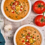 Two bowls of gazpacho soup on a rustic tile background surrounded by some of the ingredients and garnished with cherry tomatoes, cucumber and fresh parsley.