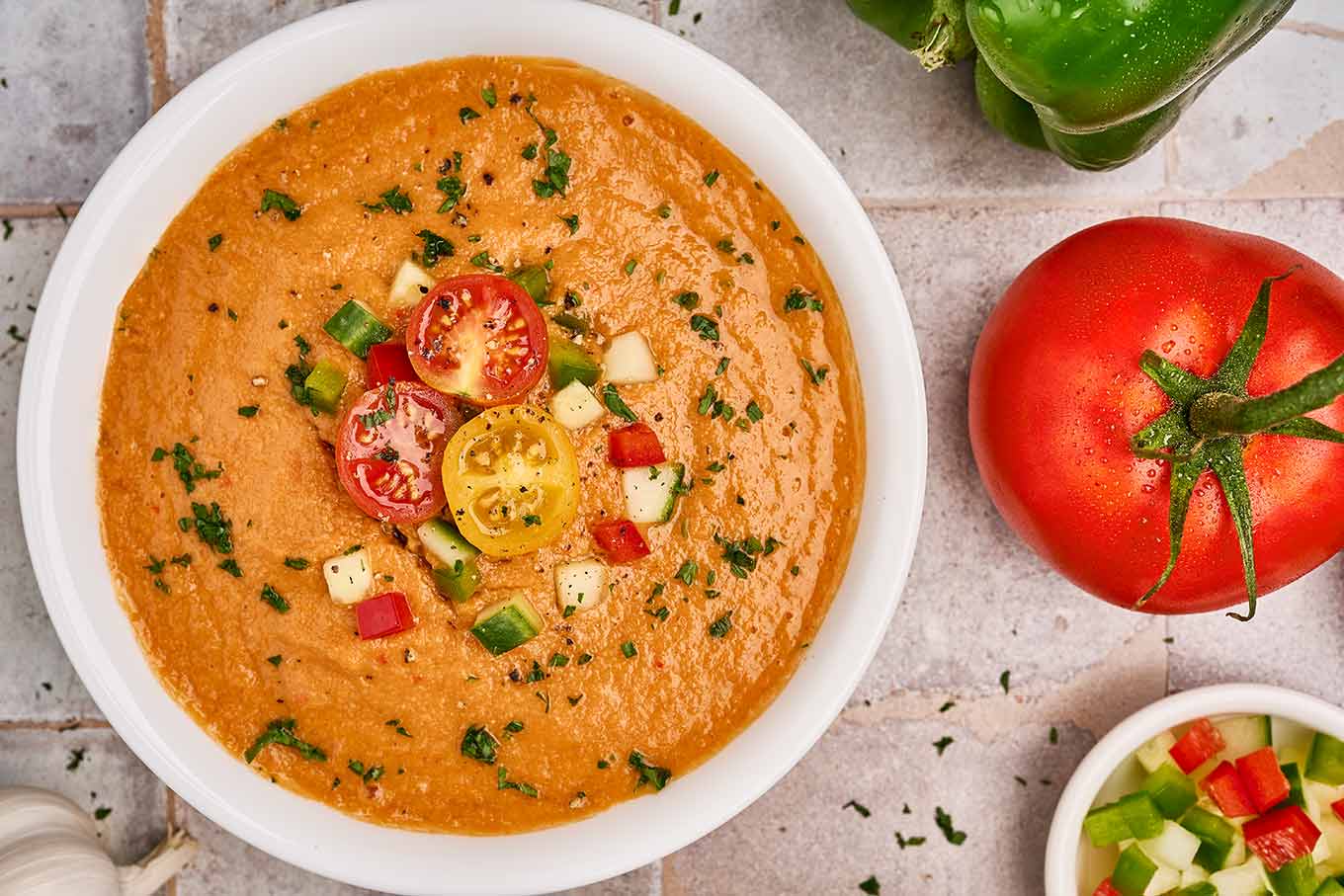 A serving of gazpacho soup in a white bowl on a tile background surrounded by fresh ingredients, and prep bowls filled with garnish.