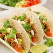 Three soft ground turkey tacos on a plate garnished with lime and cilantro, a plate filled with taco toppings in the background.
