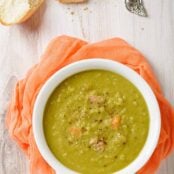 A serving of split pea soup in a white bowl with a handle on a light wooden surface with crusty bread, soup spoon, and pepper mill on the side.
