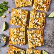 Mexican street corn flatbread cut into pieces on a baking sheet and garnished with fresh cilantro; lime wedges, garlic, and elote seasoning to the side.