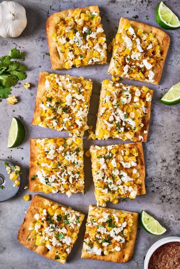 Mexican street corn flatbread cut into pieces on a baking sheet and garnished with fresh cilantro; lime wedges, garlic, and elote seasoning to the side.