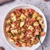 A serving bowl filled with warm potato salad with bacon dressing, serving spoon to the side, surrounded by fresh ingredients and scattered chopped chives.