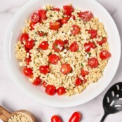 A white serving bowl filled with couscous salad next to a serving spoon, grape tomatoes, lemon wedges, and a measuring cup of pearl couscous.