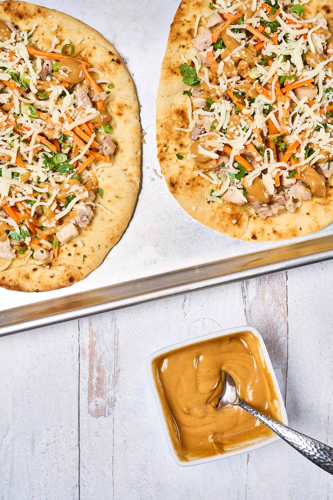 A prep photo showing two chicken flatbreads assembled and ready to be baked atop a baking sheet lined with parchment paper, peanut sauce on the side.