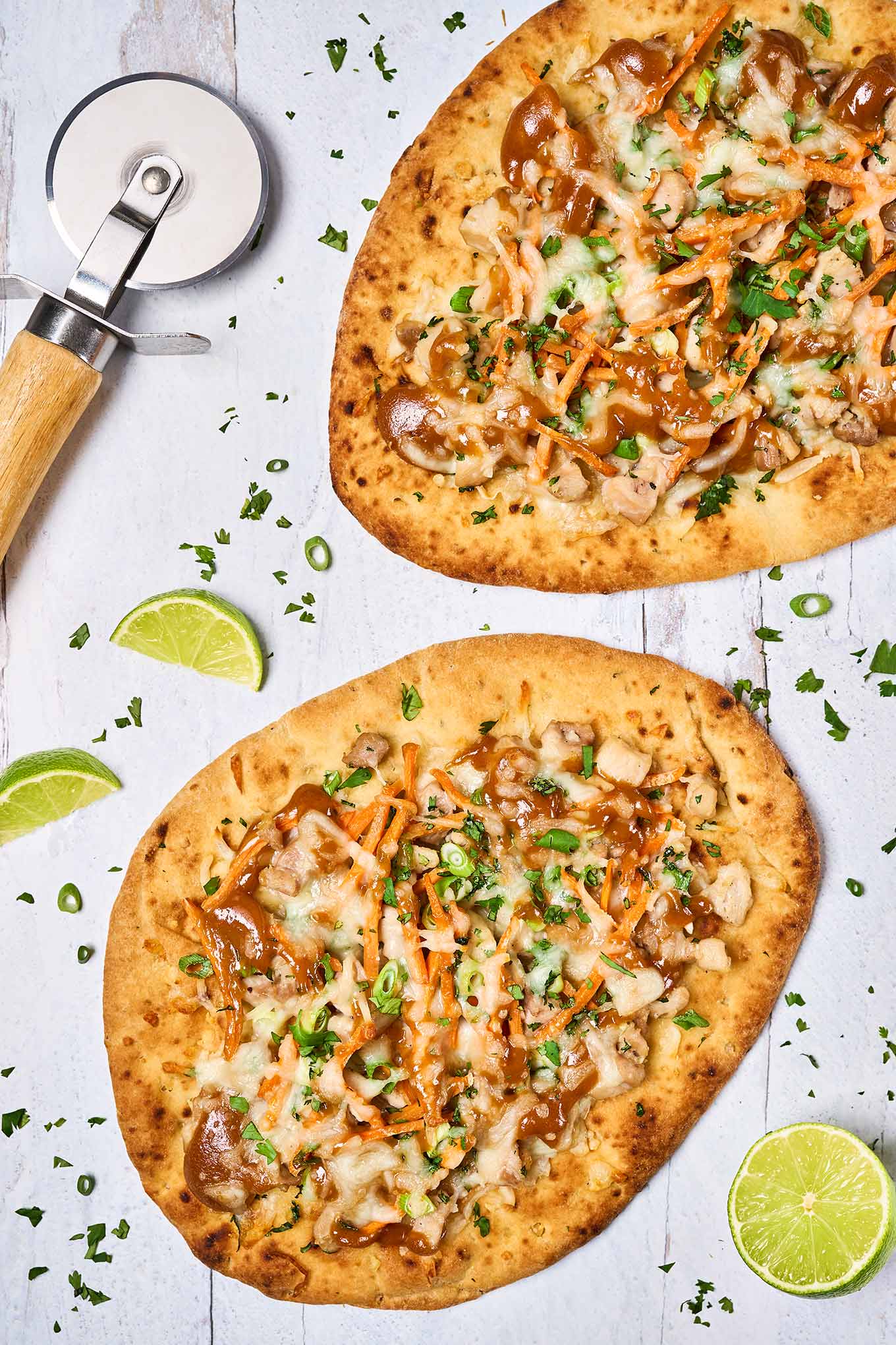 Two Thai chicken flatbread pizzas on a white wooden surface, baked and ready to be cut for serving with fresh lime wedges as garnish.